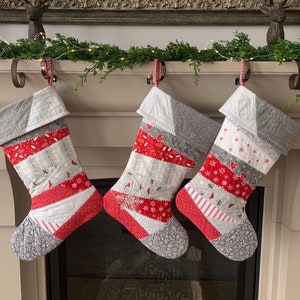 以下が含まれることがあります： 赤、白、グレーのパッチワークデザインの3つの手作りのクリスマスストッキングが暖炉の上に吊るされています。各ストッキングは、グレーのカフス、赤と白のストライプ、お祝いのパターンが特徴です。ストッキングは装飾的なフックで吊るされています。