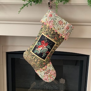 May include: A Christmas stocking with a black background and gold accents. The stocking features a red and white patterned trim and a small embroidered image of two bells with a bow. The stocking is hanging from a fireplace mantel.