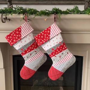 以下が含まれることがあります： 緑と光のガーランドが飾られた暖炉の棚に、赤と白のパッチワークのクリスマスストッキングが2つ吊るされています。