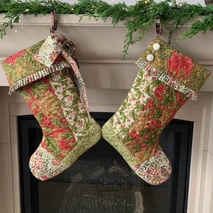以下が含まれることがあります： 赤、緑、金色の花柄のキルトクリスマスストッキング2つ。ストッキングは、緑で飾られた暖炉のマントルピースから吊り下げられています。各ストッキングには、装飾的な袖口と模様入りのつま先が付いています。