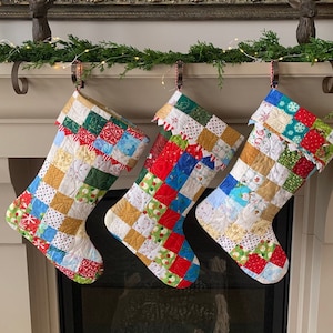 以下が含まれることがあります： 暖炉のマントルピースに、パッチワークのクリスマスストッキングが3つ吊るされています。ストッキングは、赤、緑、青、黄、白のカラフルな布の正方形で作られています。ストッキングは、白いトリムと波状の縁で飾られています。
