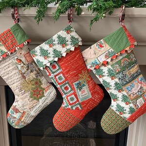 以下が含まれることがあります： パッチワークのデザインが施された赤と緑のクリスマスストッキングが3つ。ストッキングは白い暖炉の上に、緑の葉のガーランドと一緒に飾られています。