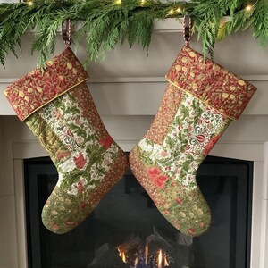 以下が含まれることがあります： 赤、緑、金色の花柄のキルトクリスマスストッキング2つ。ストッキングは暖炉の上のガーランドから吊り下げられています。ストッキングには赤と金の袖口が付いています。