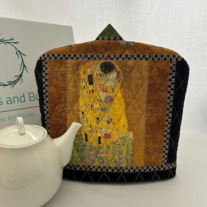 May include: A quilted tea cozy featuring Gustav Klimt's "The Kiss" artwork. The cozy has a gold, brown, and black color scheme with a diamond pattern. A white teapot is in the foreground. The cozy is designed to keep tea warm.