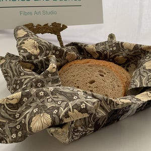 May include: A brown and beige fabric bread basket with a floral pattern. The basket is folded and contains a loaf of bread. The basket is labeled "Rambles and Bushes Fibre Art Studio".