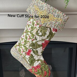 以下が含まれることがあります： 赤、緑、白の花柄のキルティングクリスマスストッキング。カフには金の刺繍と「New Cuff Style for 2026」の文字が入っています。ストッキングは暖炉の近くに吊るされています。