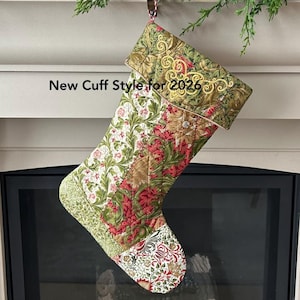 以下が含まれることがあります： 赤、緑、白の花柄のキルティングクリスマスストッキング。袖口は金色の装飾的なデザインです。「New Cuff Style for 2026」の文字がストッキングに表示されています。ストッキングは暖炉の前に吊るされています。