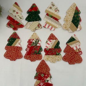 Peut inclure: Neuf décorations en tissu en forme de sapin de Noël disposées en grille. Chaque sapin est composé de bandes de tissus en coton aux motifs de fêtes rouges, verts, dorés et blancs, incluant des poinsettias, des flocons et des rayures.