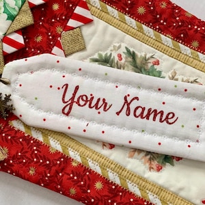 May include: White fabric ornament with red and green polka dots and the text "Your Name" embroidered in red. The ornament is attached to a gold key chain with a tassel.