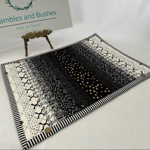 May include: A rectangular quilted placemat with a black and white striped border. The central panel features a variety of black, white, and grey patterned fabrics. The placemat has decorative buttons at the corners and a mix of geometric and floral designs.
