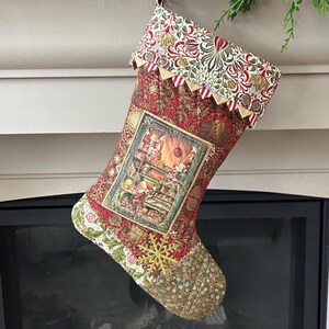May include: A decorative Christmas stocking with a detailed design. The stocking has a red background with floral patterns, a framed image, and a gold snowflake. The cuff has a contrasting floral pattern with gold trim. A festive holiday item.