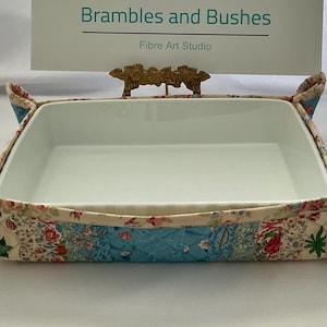 May include: A white ceramic baking dish with a floral patterned fabric cozy. The fabric features a patchwork design with blue, pink, and green floral patterns. The dish has a decorative gold-colored handle. The text "Brambles and Bushes" is visible.