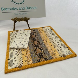May include: A quilted placemat and matching napkin set with a bee theme. The placemat features a patchwork design with various fabrics in yellow, black, and cream, and is trimmed with a yellow polka dot border. The napkin is white with a bee pattern.
