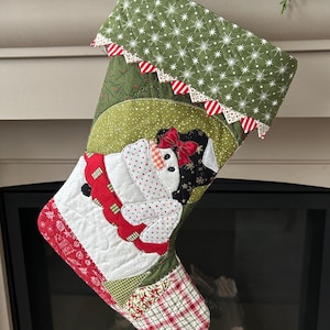 May include: A quilted Christmas stocking featuring a snowman appliqué with a red bow, wearing a polka-dot scarf. The cuff is olive green with white starbursts and red and white striped trim. The toe and heel feature red and white plaid fabric.