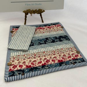 May include: A quilted placemat with a matching napkin. The placemat features a patchwork design with floral and striped fabrics in red, blue, and cream. The border is navy blue and white striped. The napkin is light blue and white striped. The placemat is secured with bronze-colored buttons.