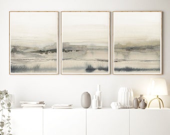 May include: Three framed abstract landscape paintings in muted tones of gray, beige, and white, hanging on a white wall above a white cabinet with a stack of books underneath.