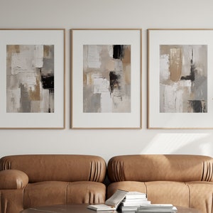 Puede incluir: Tres impresiones artísticas abstractas enmarcadas con tonos neutros de beige, gris y negro cuelgan sobre un sofá de cuero marrón. Una mesa auxiliar y una mesa de centro negras con libros y una lámpara completan la decoración moderna de la sala de estar.
