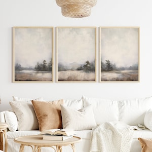 Puede incluir: Un sofá blanco con almohadas marrones y una manta blanca. El sofá está frente a una pared blanca con tres cuadros de paisajes enmarcados. Las pinturas están en tonos grises y marrones apagados.