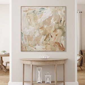 May include: A large abstract painting with a white background and brown, beige, and green brushstrokes. The painting is framed in a thin, gold frame and is hanging on a white wall above a wooden console table.