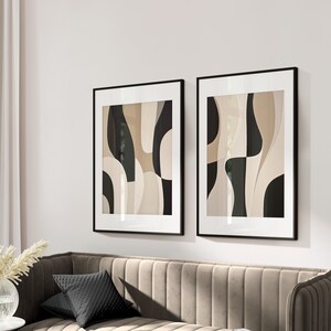 Op de afbeelding: Twee zwarte ingelijste abstracte kunstprints met beige en zwarte geometrische vormen. De prints hangen aan een witte muur boven een beige bank met zwarte kussens.