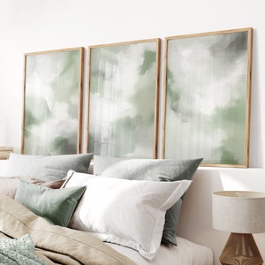 Könnte beinhalten: Ein Schlafzimmer mit einem Bett mit weißer und brauner Bettwäsche und einer grünen Wolldecke. An der Wand über dem Bett hängen drei gerahmte abstrakte Gemälde in Grün- und Weißtönen. Eine Holzlampe mit weißem Schirm steht auf einem Korbtisch neben dem Bett.