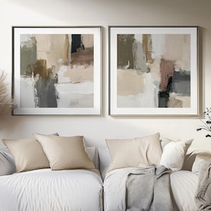 May include: Two framed abstract paintings in neutral tones hang above a white sofa. The artwork features brushstrokes of beige, grey, black, and white. The sofa has beige cushions and a grey throw.
