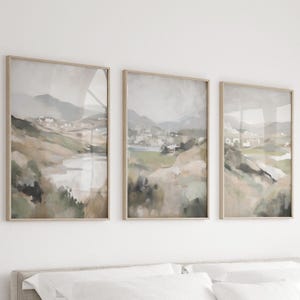 Puede incluir: Un conjunto de tres pinturas de paisajes enmarcadas en tonos apagados de verde, marrón y gris. Las pinturas representan una colina ondulada con un pequeño pueblo en la distancia.