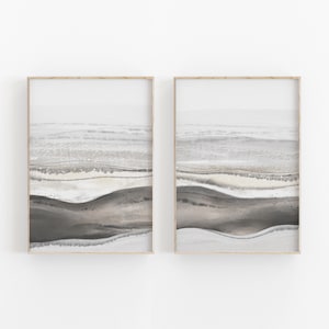 May include: Two framed abstract watercolor paintings with a minimalist style. The paintings feature a layered landscape with shades of gray, brown, and white.