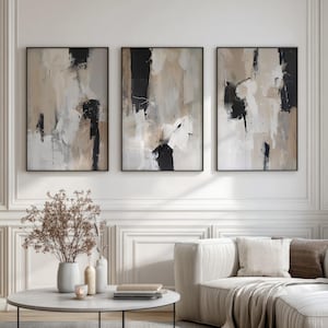 Puede incluir: Tres obras de arte abstractas enmarcadas. Cada pieza presenta una mezcla de pinceladas negras, beige y blancas. La obra de arte se exhibe sobre un sofá blanco y una mesa de centro redonda con un jarrón de flores secas.