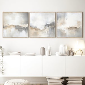 Puede incluir: Tres pinturas abstractas enmarcadas con tonos neutros de gris, beige y blanco. La obra de arte tiene una composición similar a un paisaje con una apariencia texturizada y mezclada. Los marcos son de color madera clara. Las pinturas se exhiben sobre un mueble blanco.