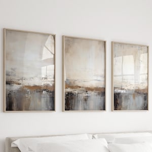Puede incluir: Un conjunto de tres pinturas abstractas de paisajes en tonos apagados de marrón, gris y blanco. Las pinturas están enmarcadas en un marco de madera claro.