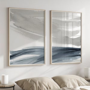 May include: Two framed abstract watercolor paintings with a gray and blue color scheme. The paintings are hung on a white wall above a bed with beige linens.