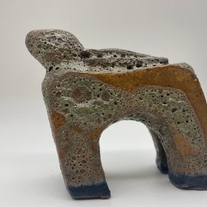 May include: A ceramic sculpture of an abstract animal with a textured surface. The sculpture is brown, grey, and yellow with a blue base.