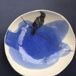 Könnte beinhalten: Eine weiße Keramikschale mit einem blauen und weißen Design. Die Schale hat einen Riss und eine goldene Linie verläuft durch sie. Ein schwarzer Rabe ist auf die Schale gemalt.