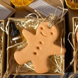 Könnte beinhalten: Ein kleiner Keramik-Lebkuchenmann-Anhänger in einer Schachtel mit zerknülltem Papier. Eine kleine Flasche mit Zimt-Ätherischem Öl ist enthalten. Die Schachtel hat ein Etikett mit der Aufschrift "Einfach 1 bis 2 Tropfen ätherisches Öl zu Ihrer porösen Keramikdekoration hinzufügen und an Ihren Weihnachtsbaum, Heizkörper, Ihr Auto oder Ihren Wunschort hängen. Zum Auffrischen einfach noch einen Tropfen oder zwei hinzufügen."