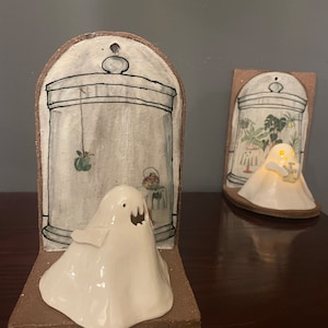 Peut inclure: Deux serre-livres en céramique en forme de fantômes avec un glaçage blanc. Les serre-livres sont en forme de fantômes avec une petite ouverture pour une lumière. Les serre-livres sont décorés d'une illustration en noir et blanc d'une maison avec une fenêtre et une porte. Les serre-livres sont posés sur une surface en bois.