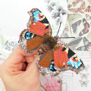 May include: A handcrafted butterfly brooch with intricate embroidery and sequin detailing. The butterfly features vibrant orange, red, blue, and black wings, with a brown body. The wings are adorned with silver sequins. The brooch is a unique accessory.