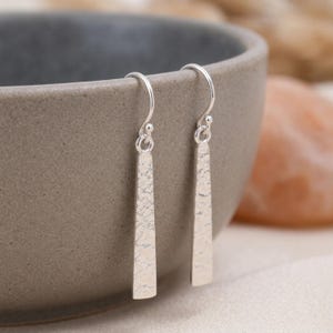 May include: A pair of silver drop earrings featuring elongated, tapered bar pendants with a textured, hammered finish. Each earring hangs from a simple curved silver ear wire, showcasing a metallic sheen and a distinct, uneven surface pattern.