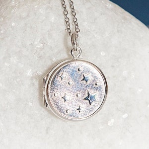 Puede incluir: Collar con colgante plateado y cadena fina. El colgante redondo presenta un dise&ntilde;o celestial de estrellas y puntos. El colgante est&aacute; cerrado, lo que sugiere que puede contener una peque&ntilde;a foto o recuerdo. El collar se muestra sobre una superficie blanca.