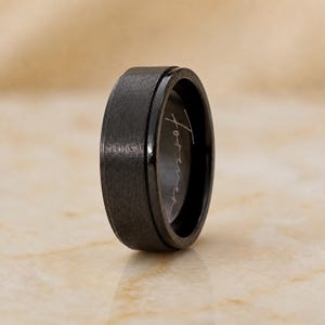 May include: A black ring with a brushed, textured outer surface and a smooth, polished inner band. The word "forever" is engraved in a cursive script on the interior surface of the ring, which is positioned upright to show both the exterior and interior.