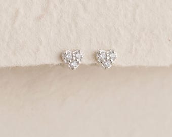 May include: A pair of silver stud earrings, each shaped like a heart and set with three small, clear, sparkling stones. The earrings feature a polished metallic finish and a delicate, symmetrical design, showcasing a classic heart silhouette.