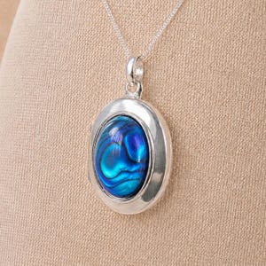 May include: An oval locket pendant featuring a polished silver frame and a central iridescent abalone shell inlay with vibrant blue and green swirling patterns, suspended from a delicate silver chain link necklace.