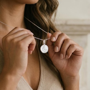 Op de afbeelding: Een zilverkleurige ketting met een ronde hanger waarop een figuur met een staf is afgebeeld. De hanger hangt aan een fijne zilveren ketting en heeft een gepolijste metalen afwerking met een gedetailleerd, gegraveerd ontwerp op de munt.