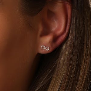 Peut inclure: Gros plan d'une boucle d'oreille en forme de symbole infini argenté. La boucle d'oreille est petite et détaillée, avec une surface texturée. La boucle d'oreille est portée sur une oreille, l'accent étant mis sur le bijou.