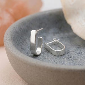 May include: A pair of silver hoop earrings with a smooth, polished finish and a distinct U-shaped design. Each earring features a straight post and a secure backing mechanism, showcasing a clean, reflective metallic surface and a simple, open structure.