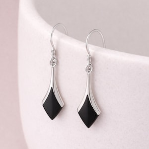 May include: A pair of silver drop earrings with a geometric design. Each earring has a black, diamond-shaped inlay framed by a silver border. The earrings hang from a curved hook.