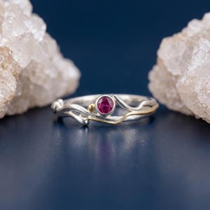 May include: A silver ring featuring a small, round, bezel-set pink gemstone and a tiny gold bead accent. The band has an organic, wavy, and textured design with a thin gold-toned wire detail integrated into the silver metal structure.