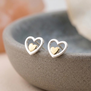 May include: A pair of stud earrings featuring a silver-toned open heart outline with a smaller, solid gold-toned heart centred within each frame. The earrings have a polished metallic finish and include a post backing for secure attachment.