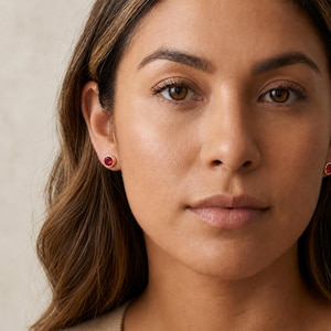 Puede incluir: Un par de pendientes de botón con piedras preciosas redondas de color rojo intenso engastadas en un sencillo marco de metal dorado. Los pendientes son pequeños y circulares, aportando un sutil toque de color al lóbulo de la oreja.