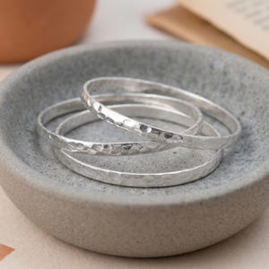 May include: Three silver-coloured stacking rings with a hammered, textured finish. The rings are thin, circular bands with a reflective metallic surface, arranged in a nested formation within a small, grey, circular stone-like bowl.